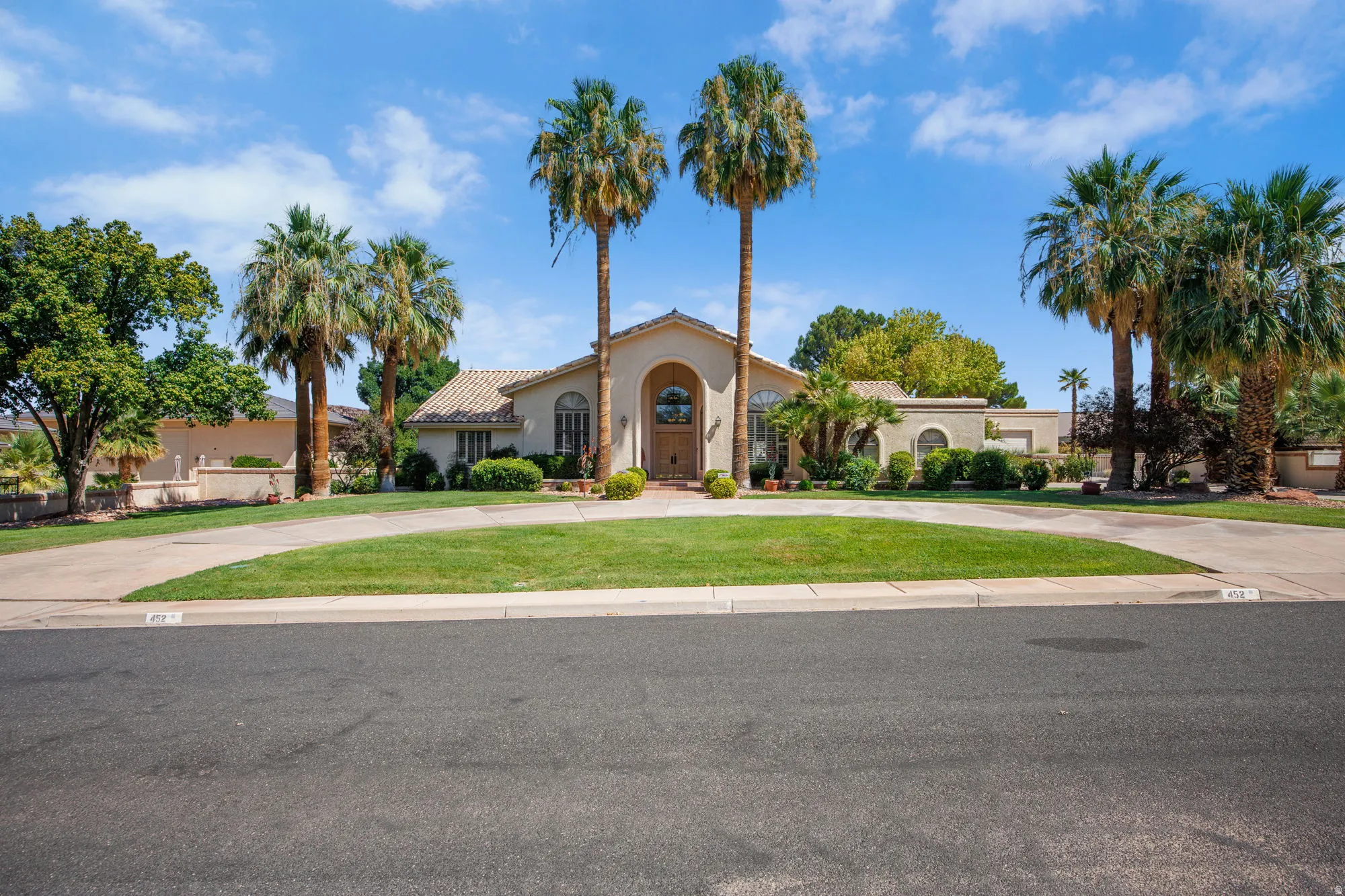 Stucco exterior with arched entry, terracotta roof tiles, mature palm trees, and a circular driveway