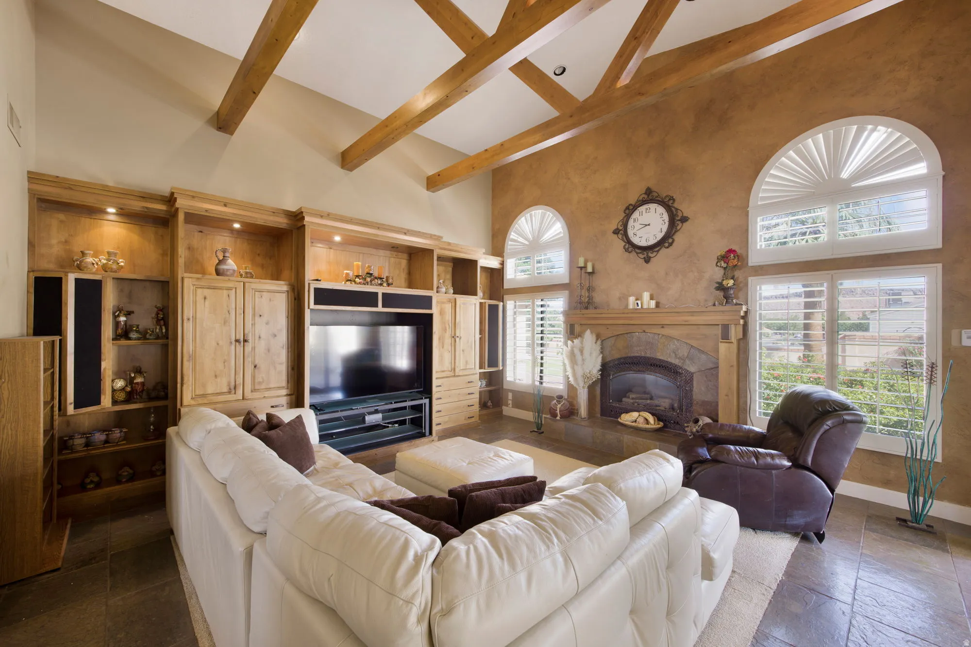Vaulted ceiling with exposed wood beams, built-in entertainment center, stone-clad fireplace with mantel, arched windows with plantation shutters, and tile flooring