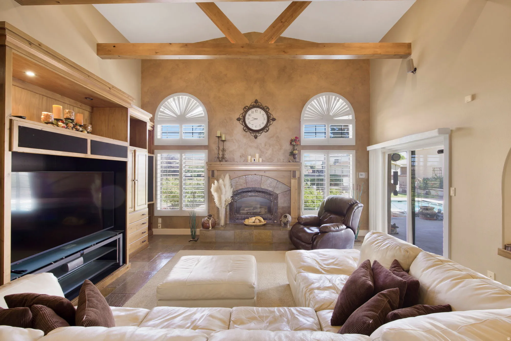 Sophisticated great room showcasing exposed wood beam ceiling architecture, dual arched transom windows with plantation shutters, and a linear fireplace with stone surround
