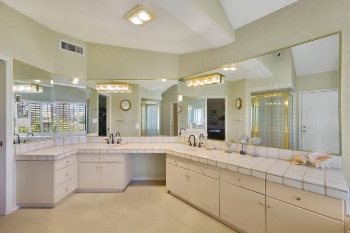 Luxurious primary bathroom suite featuring dual vanities with ample storage, extensive mirror wall with integrated lighting, tile countertops, and abundant natural light from corner window placement