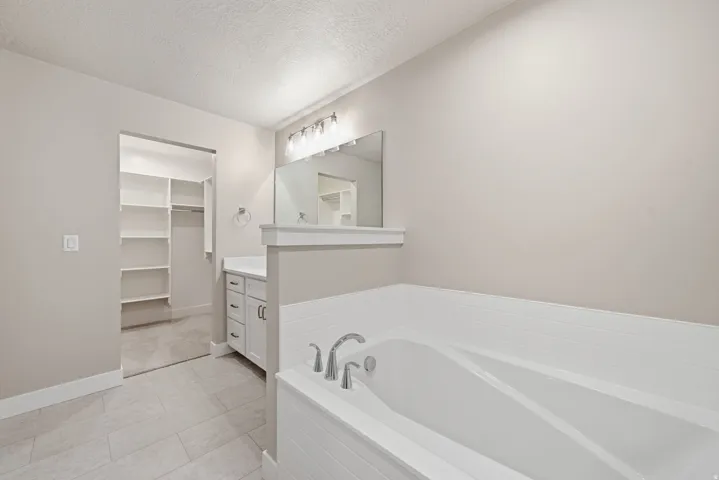 Soaking tub with tile surround, white vanity with storage, vanity mirror with overhead lighting, tile flooring, and built-in closet shelving