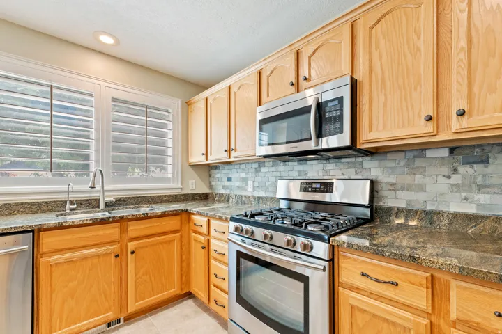 Wood-finish cabinetry, stainless steel appliances, grey stone countertops, and a tile backsplash