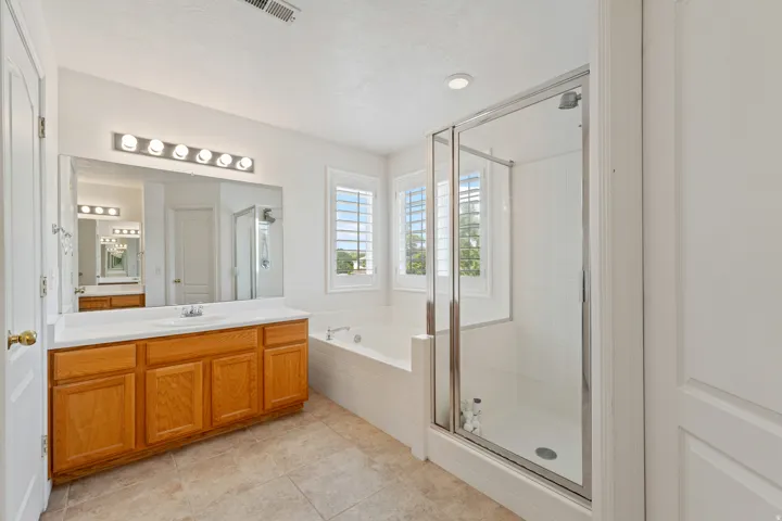 Vanity with wood-finish cabinetry, built-in soaking tub, glass-enclosed shower, plantation shutters, and large wall-mounted mirror