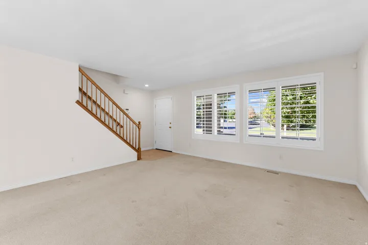 Neutral carpet flooring, white walls, wood staircase with balusters, multiple windows with white shutters, and a white entry door