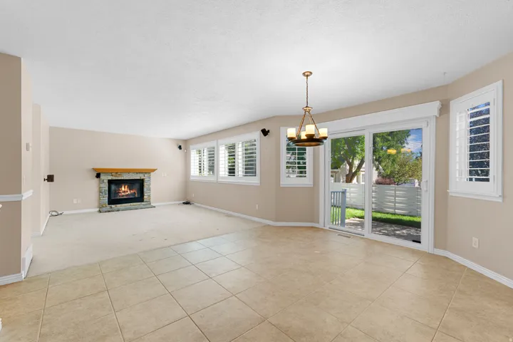 Stone-clad fireplace with a wood mantle, sliding glass patio door, tile flooring, carpeted area, and multiple windows with plantation shutters