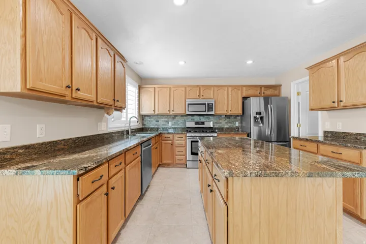 Kitchen featuring warm-toned cabinetry, granite countertops, a central island, stainless steel appliances, and a tiled backsplash