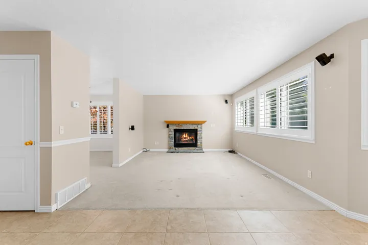 Stone fireplace with wood mantel, multiple windows with white plantation shutters, and a mixture of tile and carpet flooring
