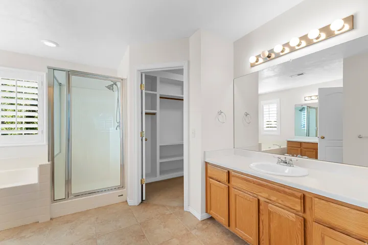 Dual sink vanity with wood cabinetry, tiled flooring, glass-enclosed shower, built-in bathtub, and a walk-in closet with shelving
