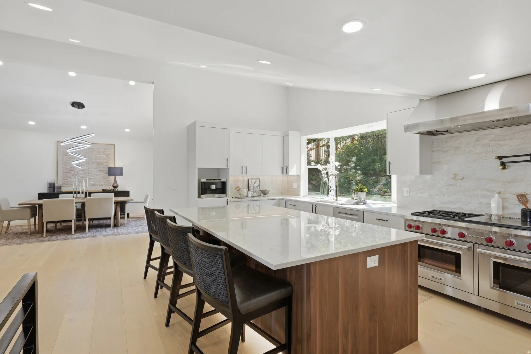 Open-concept kitchen with a large central island, light wood-finish flooring, recessed lighting, and a stainless steel range with matching hood