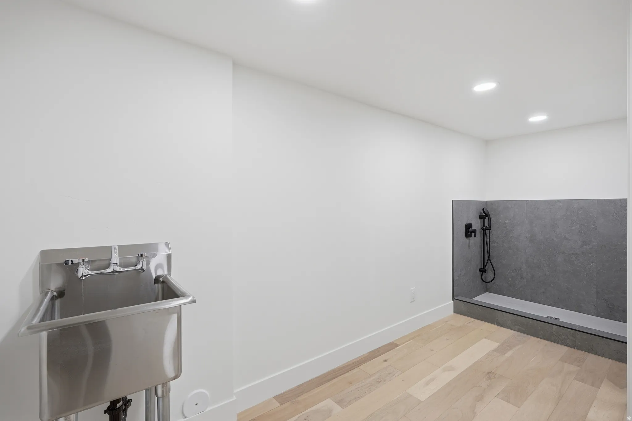Stainless steel utility sink, recessed ceiling lighting, light wood-finish flooring, and a dedicated wash station with dark-toned wall tile and matte black fixtures