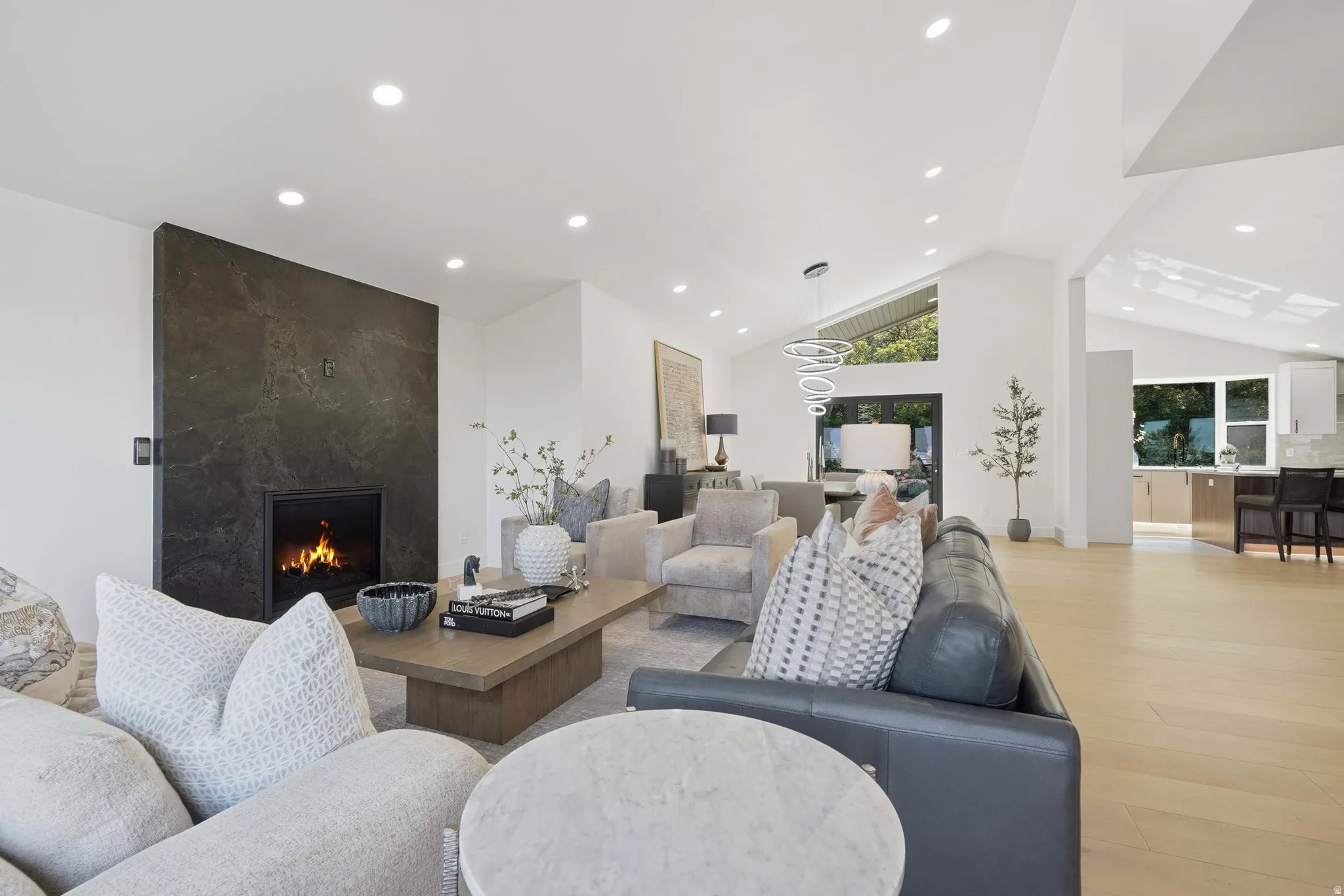 Contemporary open-concept living space featuring a dark-finished fireplace surround with integrated hearth, light wood-finish flooring throughout, recessed ceiling lighting, and expansive sightlines to adjacent dining and kitchen areas