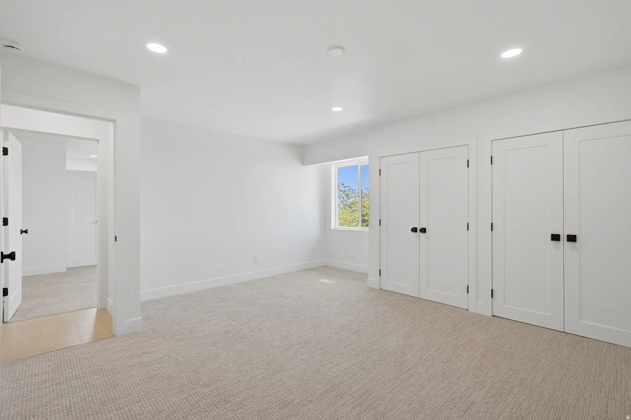 Recessed lighting, light-toned carpeting, white walls, multiple paneled doors with matte black hardware, and a window with exterior visibility