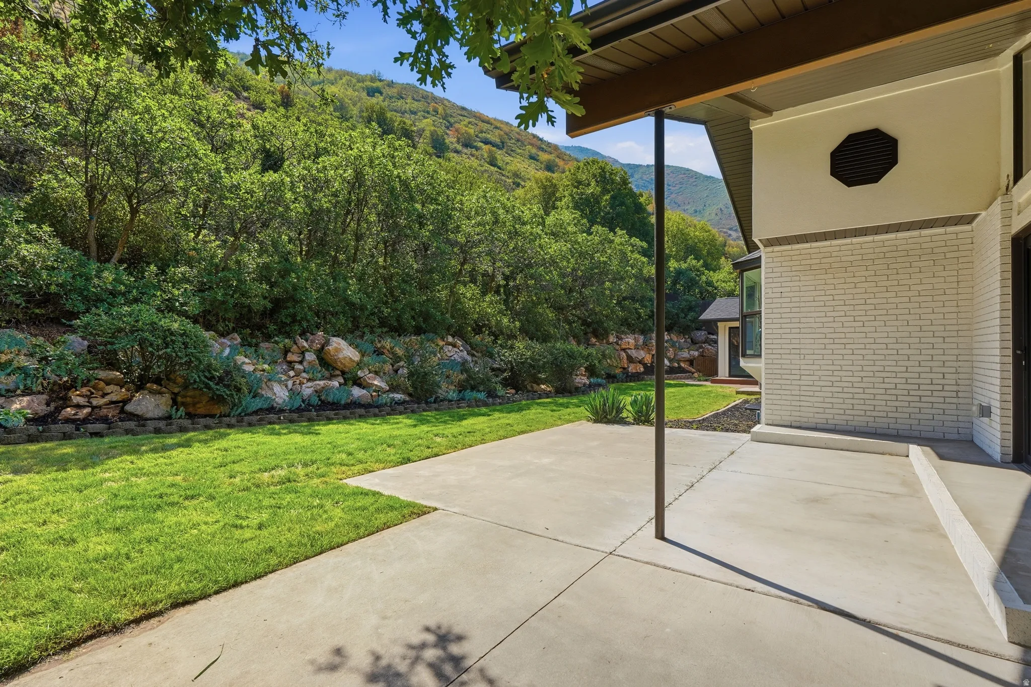 Expansive concrete patio with a covered section, bordered by a manicured lawn and a rock retaining wall with established plantings
