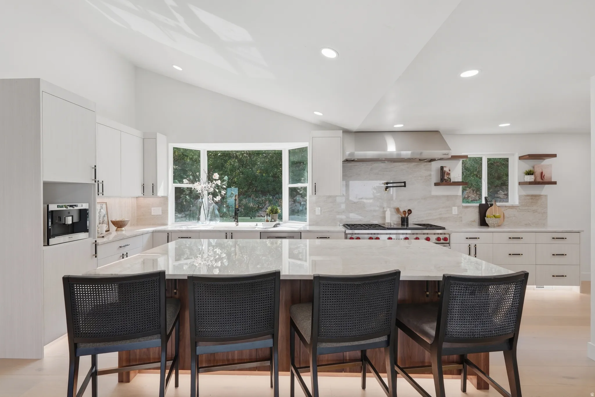 Vaulted ceiling kitchen featuring an expansive central island, custom cabinetry with integrated appliances, a bay window, and a professional-grade range with a stainless steel vent hood