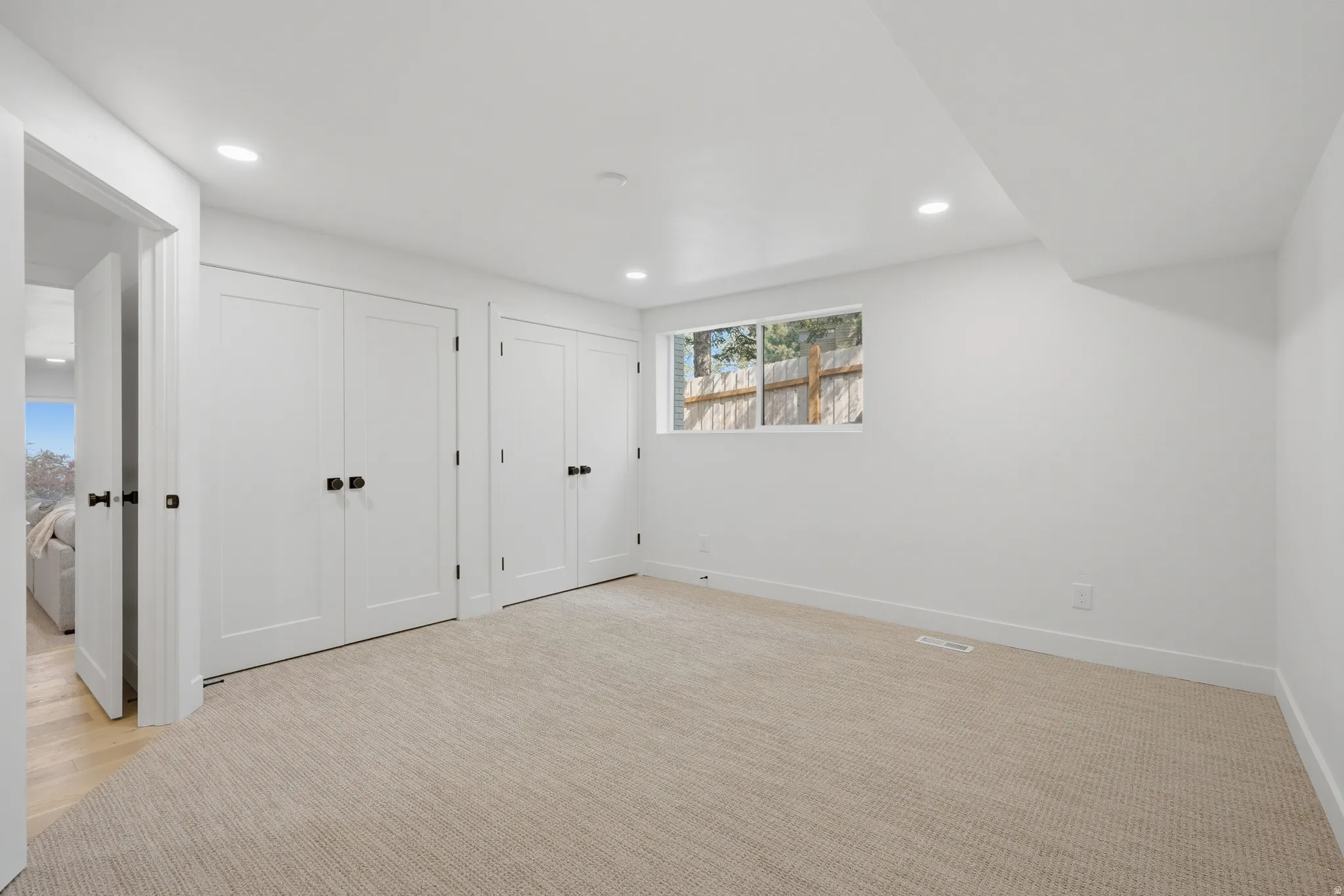 Recessed lighting, light-toned carpeting, white walls, and a window with a wooden fence background