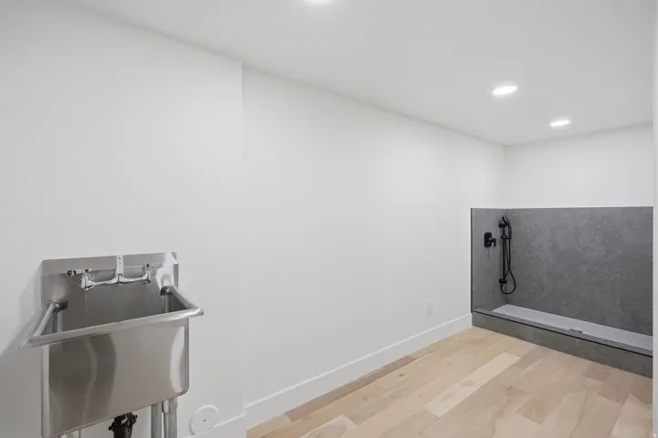 Stainless steel utility sink, recessed ceiling lighting, light wood-finish flooring, and a dedicated wash station with dark-toned wall tile and matte black fixtures