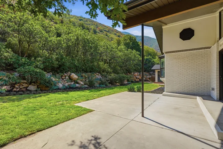 Expansive concrete patio with a covered section, bordered by a manicured lawn and a rock retaining wall with established plantings