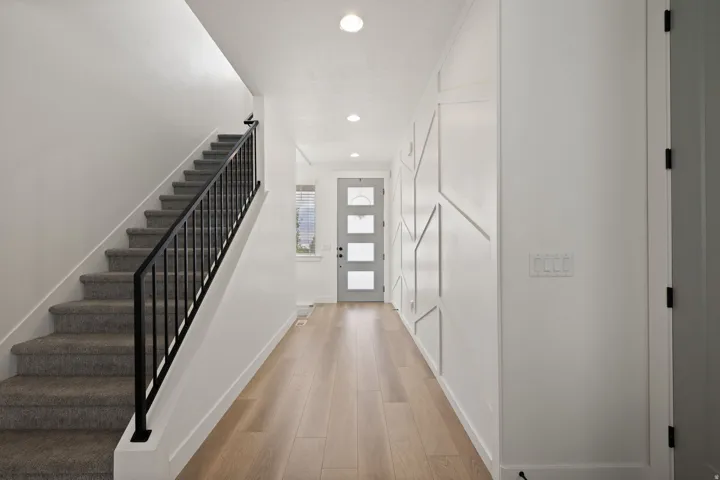 Wood-finish flooring, staircase with carpeted treads, black metal railing, decorative wall paneling, and a contemporary entry door with frosted glass panels