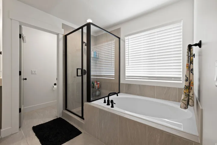 Built-in soaking tub with black fixtures, enclosed shower with black frame, tile flooring, and integrated window