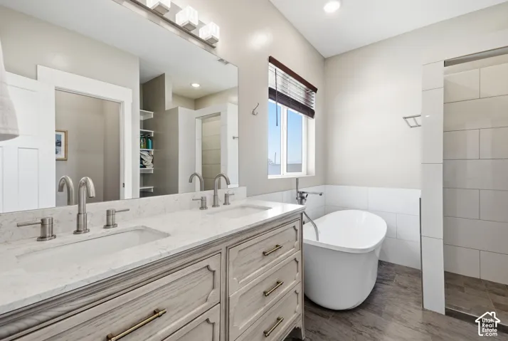 Dual vanity with stone countertop, freestanding soaking tub, separate shower enclosure with tile surround, and wood-look flooring