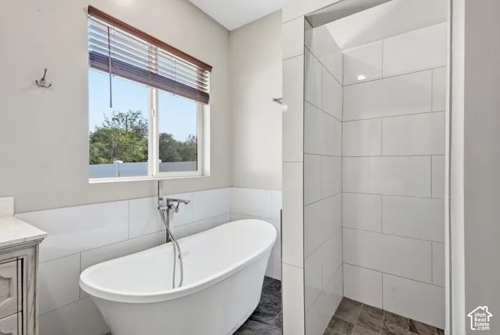 Freestanding soaking tub with a floor-mounted faucet, complemented by a tiled shower enclosure with a dark-toned floor, a vanity with a light-colored countertop, and a window with wood-finish blinds