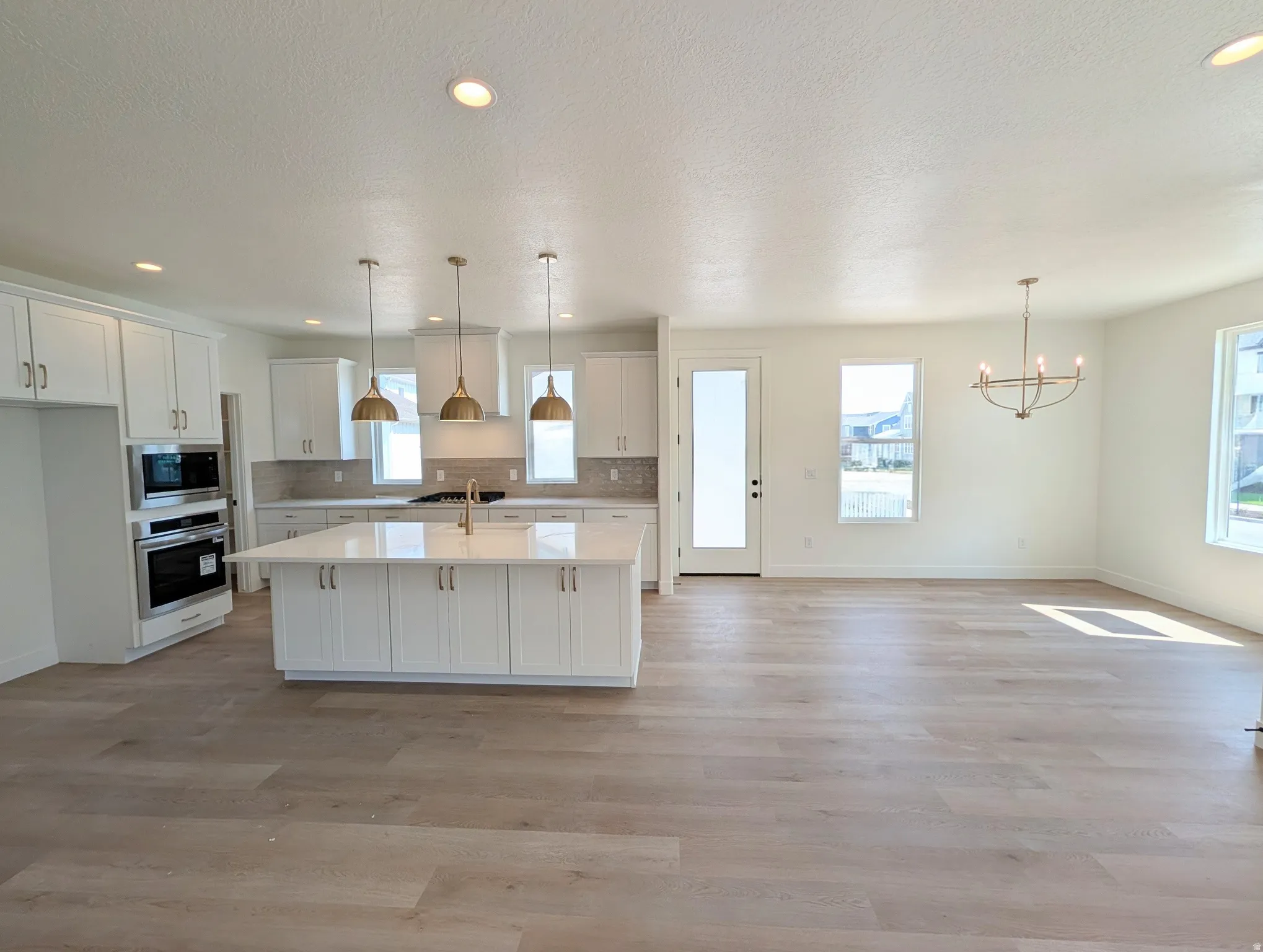 Open-concept living space with wood-finish flooring, white cabinetry, stainless steel double ovens, a large kitchen island with pendant lighting, and a modern chandelier
