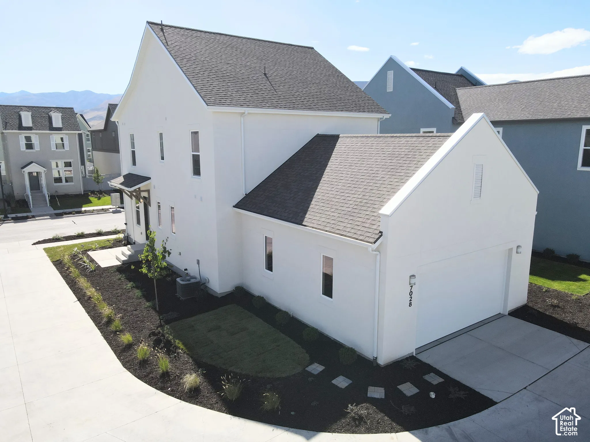 White stucco exterior, dark shingle roof, attached garage, concrete driveway, landscaped yard with planted beds