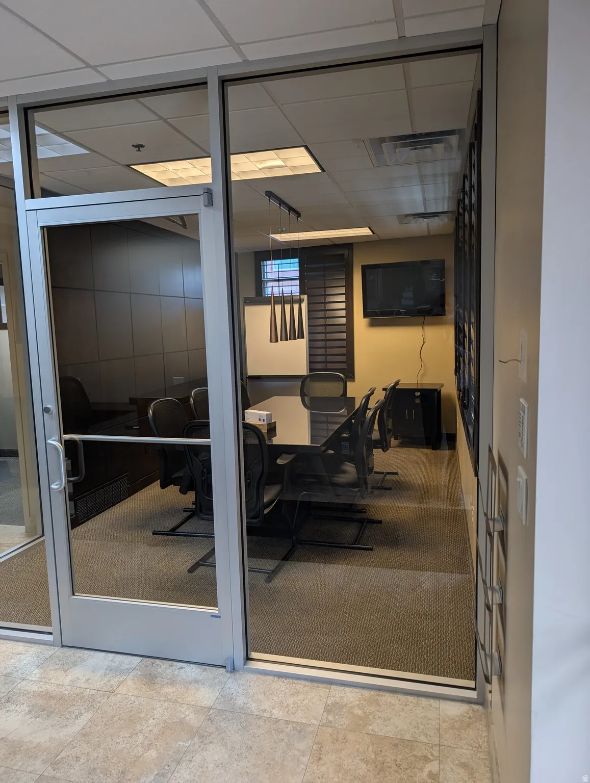 Glass-paneled entry door, commercial-grade carpet, suspended ceiling with recessed lighting, and wall-mounted screen