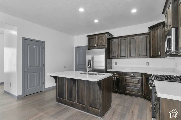 Kitchen featuring wood-finish flooring, dark wood cabinetry, stainless steel appliances, a center island with sink, and light-toned countertops