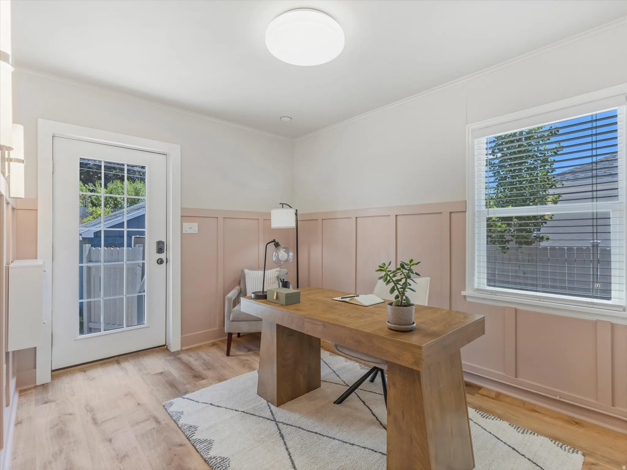 Bright home office featuring warm wood-finish flooring, wainscoting with soft pink tones, and abundant natural light from dual windows
