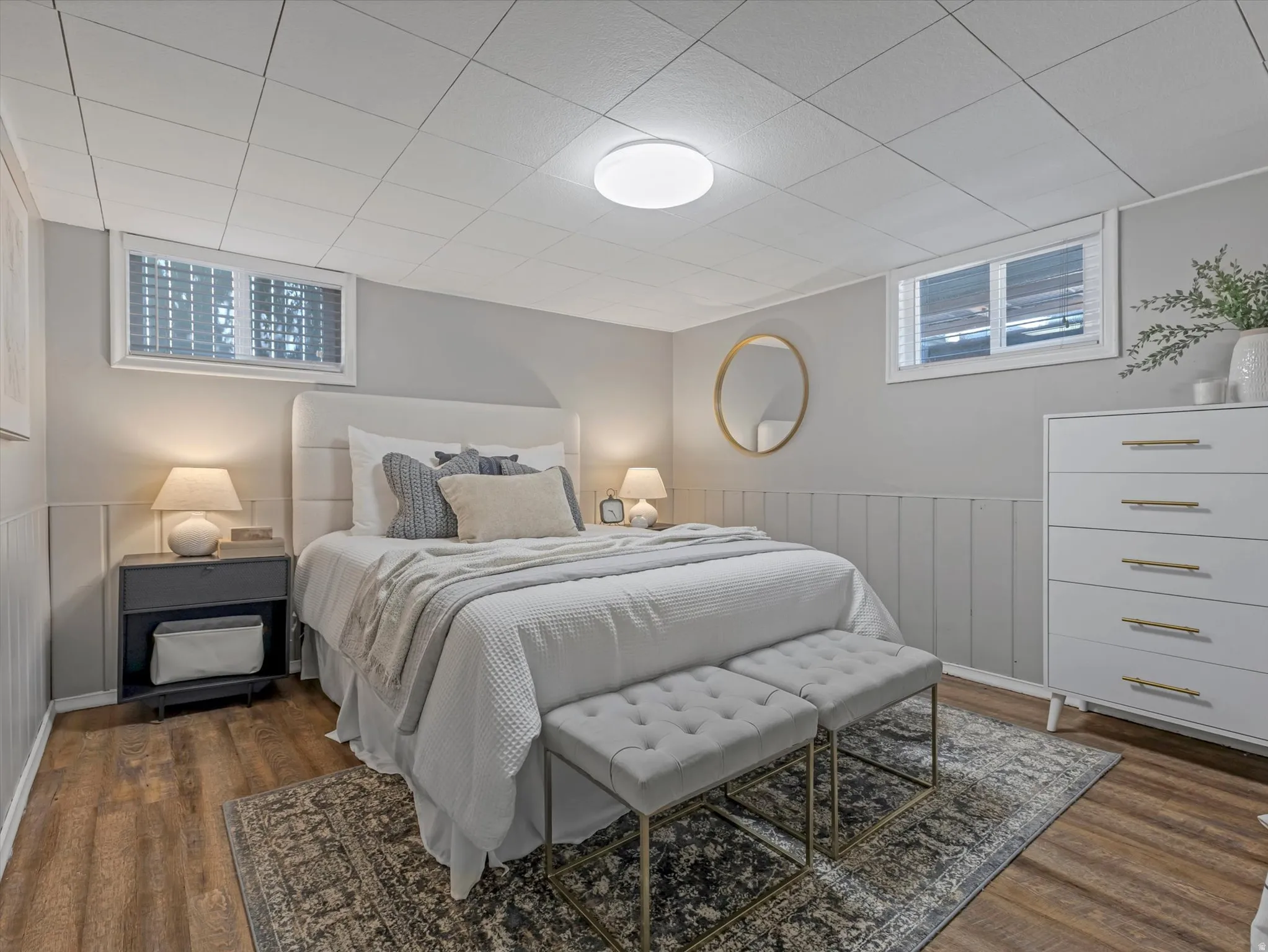 Light gray walls with vertical paneling, wood-finish flooring, recessed ceiling with a flush-mount light fixture, and two windows with blinds