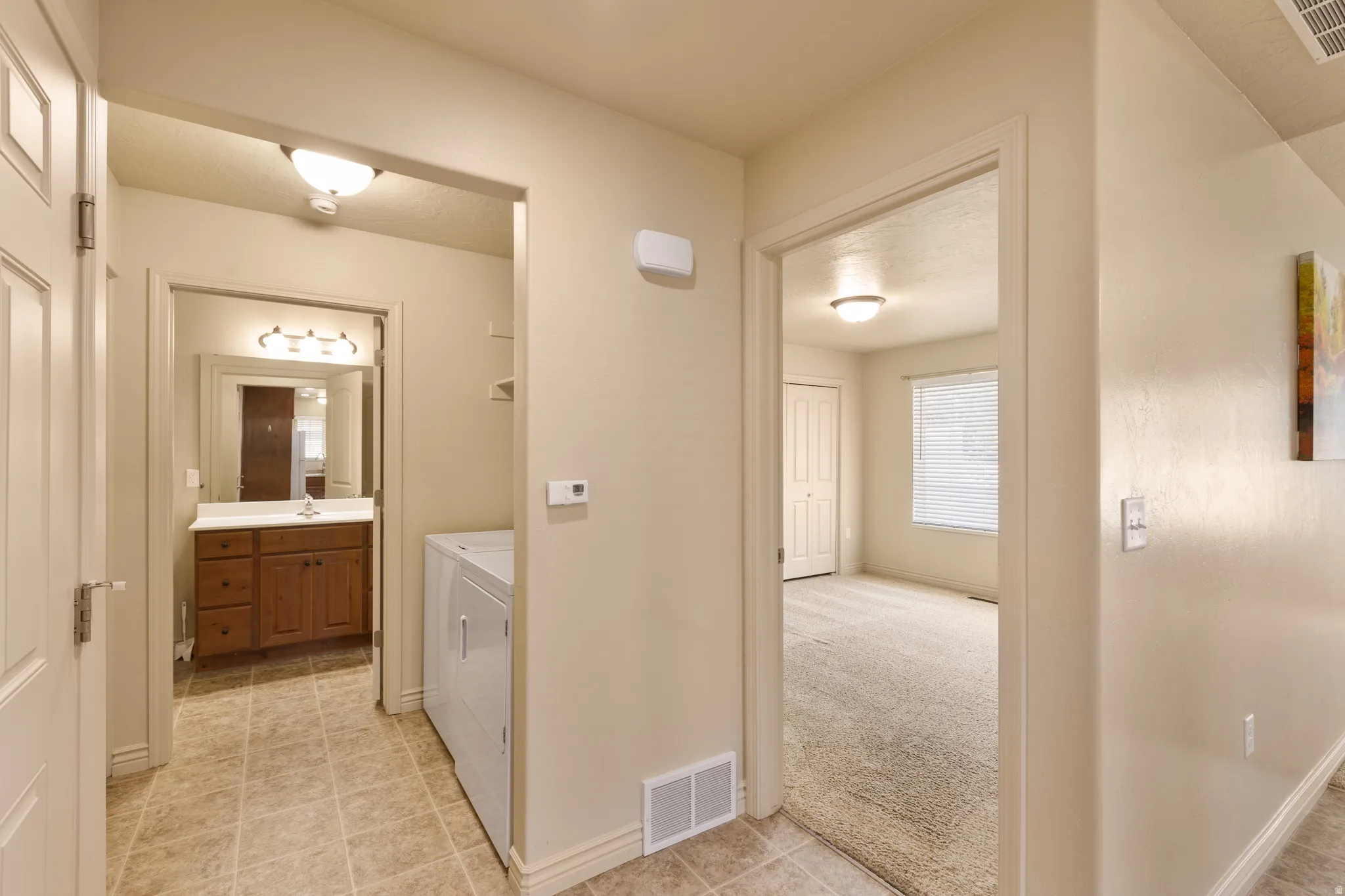 Hallway with tiled flooring, leading to a bathroom with a wood vanity and a room with carpeted flooring and a window with blinds