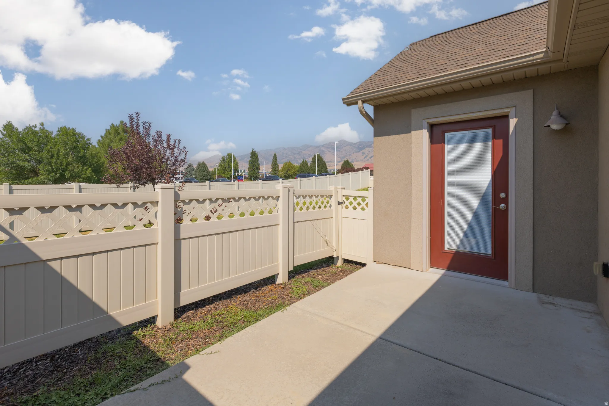 Private patio area featuring a concrete surface, red-paneled door with glass insert, exterior wall sconce, and lattice-top fencing
