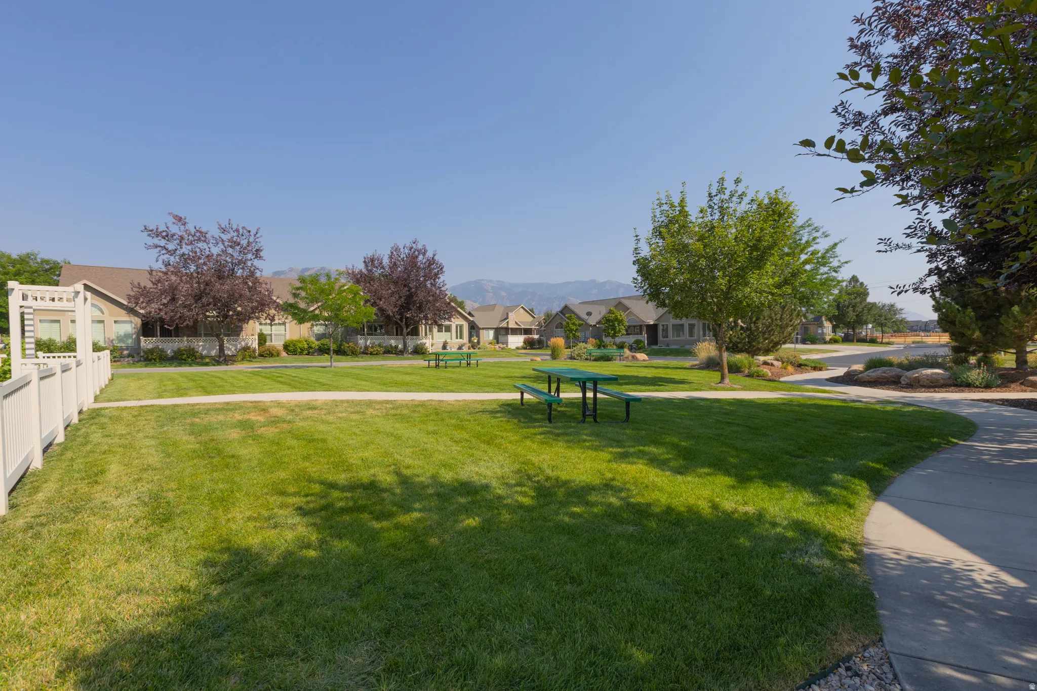 Expansive green space featuring mature trees, a paved walking path, and permanent picnic tables