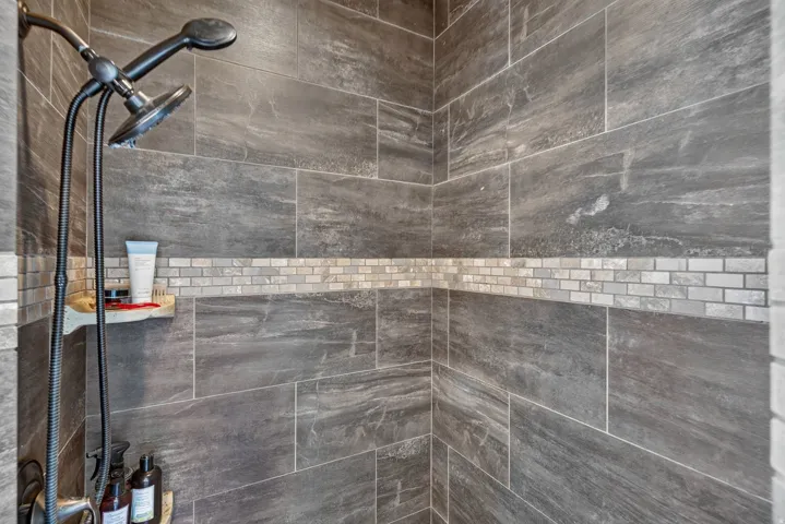 Wall-mounted shower system with a handheld showerhead, large format tile walls, mosaic tile accent band, and an integrated shower shelf