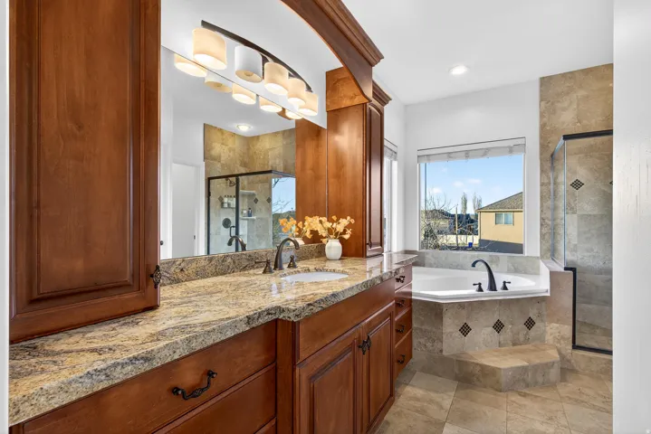 Built-in cabinetry with ample storage, a large vanity mirror with overhead lighting, an integrated soaking tub, and a glass-enclosed shower