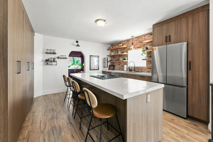 Kitchen featuring wood-finish cabinetry, a large kitchen island with a light-toned countertop, stainless steel refrigerator, and exposed brick backsplash