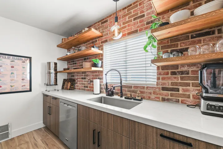 Brick accent wall, wood-finish open shelving, stainless steel sink, contemporary cabinetry, and light-toned countertops