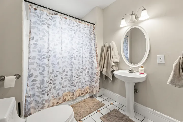 Pedestal sink with an oval mirror, tile flooring, and a shower with a grab bar