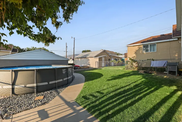 Expansive backyard featuring a manicured lawn, an above-ground pool with surrounding gravel, a concrete pathway, and a detached building