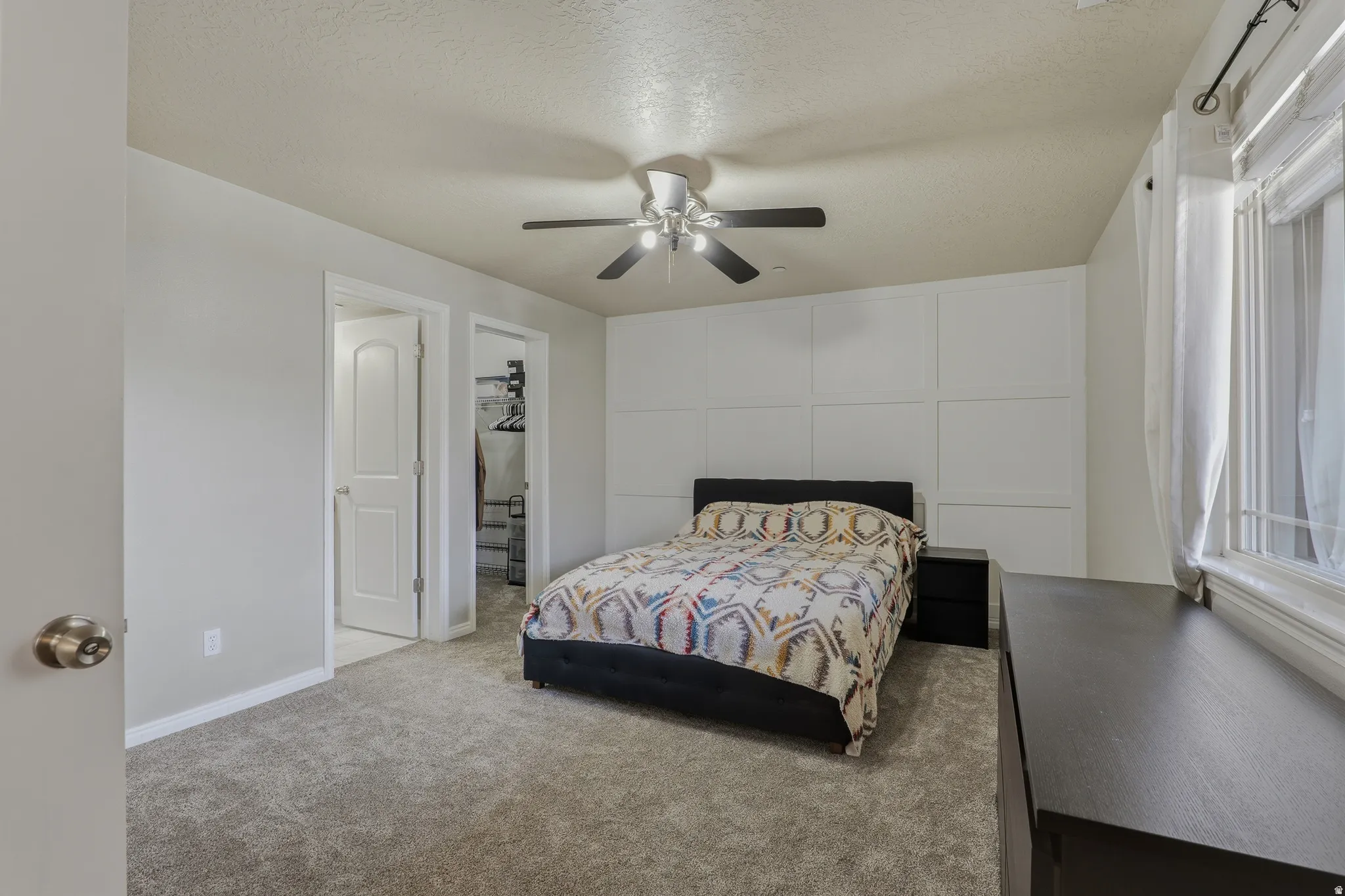 Textured wall paneling, ceiling fan with integrated lighting, carpeted flooring, and a window with curtain rod