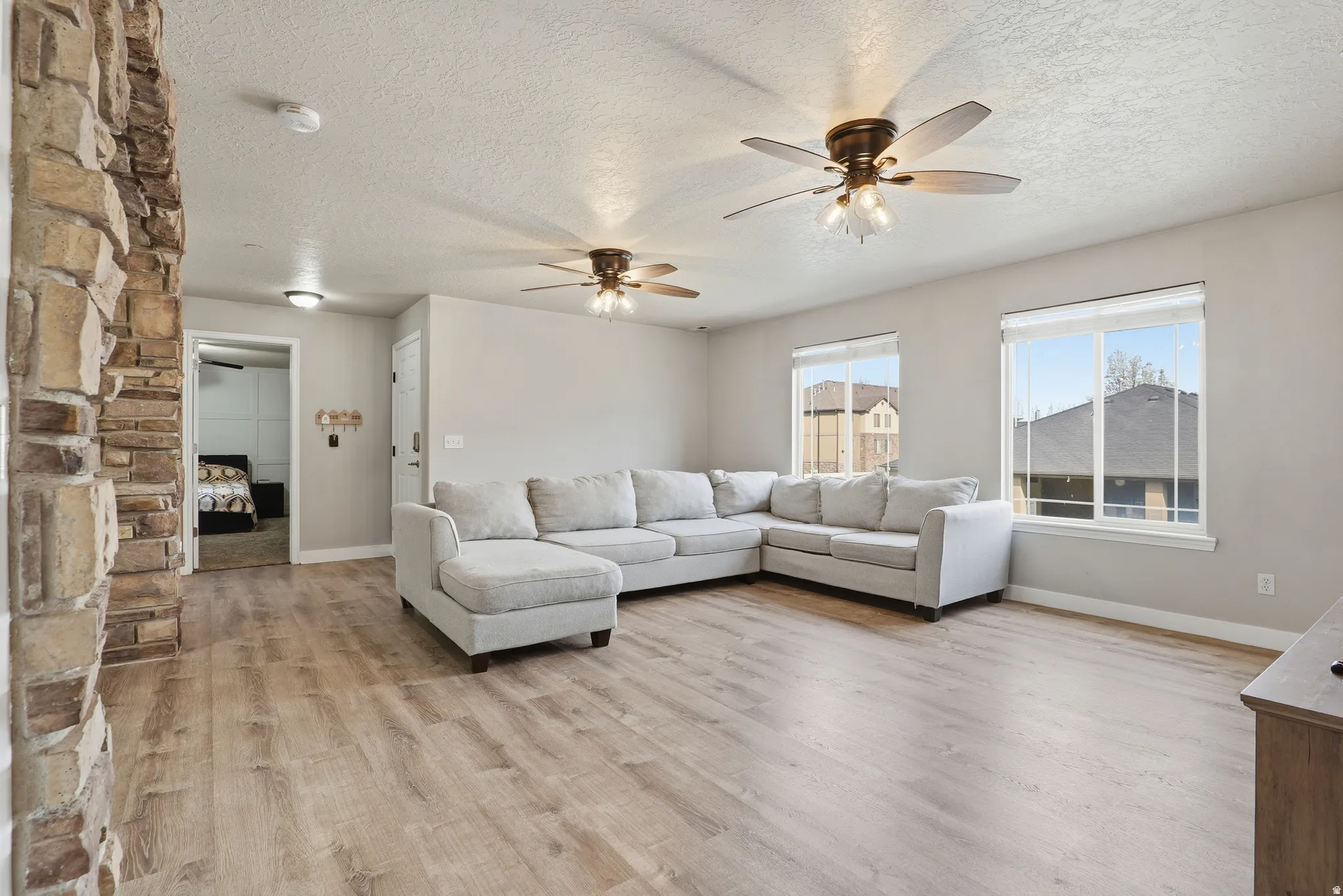 Spacious living area showcasing wood-finish flooring, a light gray upholstered sectional with chaise, dual ceiling fans with integrated lighting, large windows with city views, and a striking stacked-stone fireplace accent wall