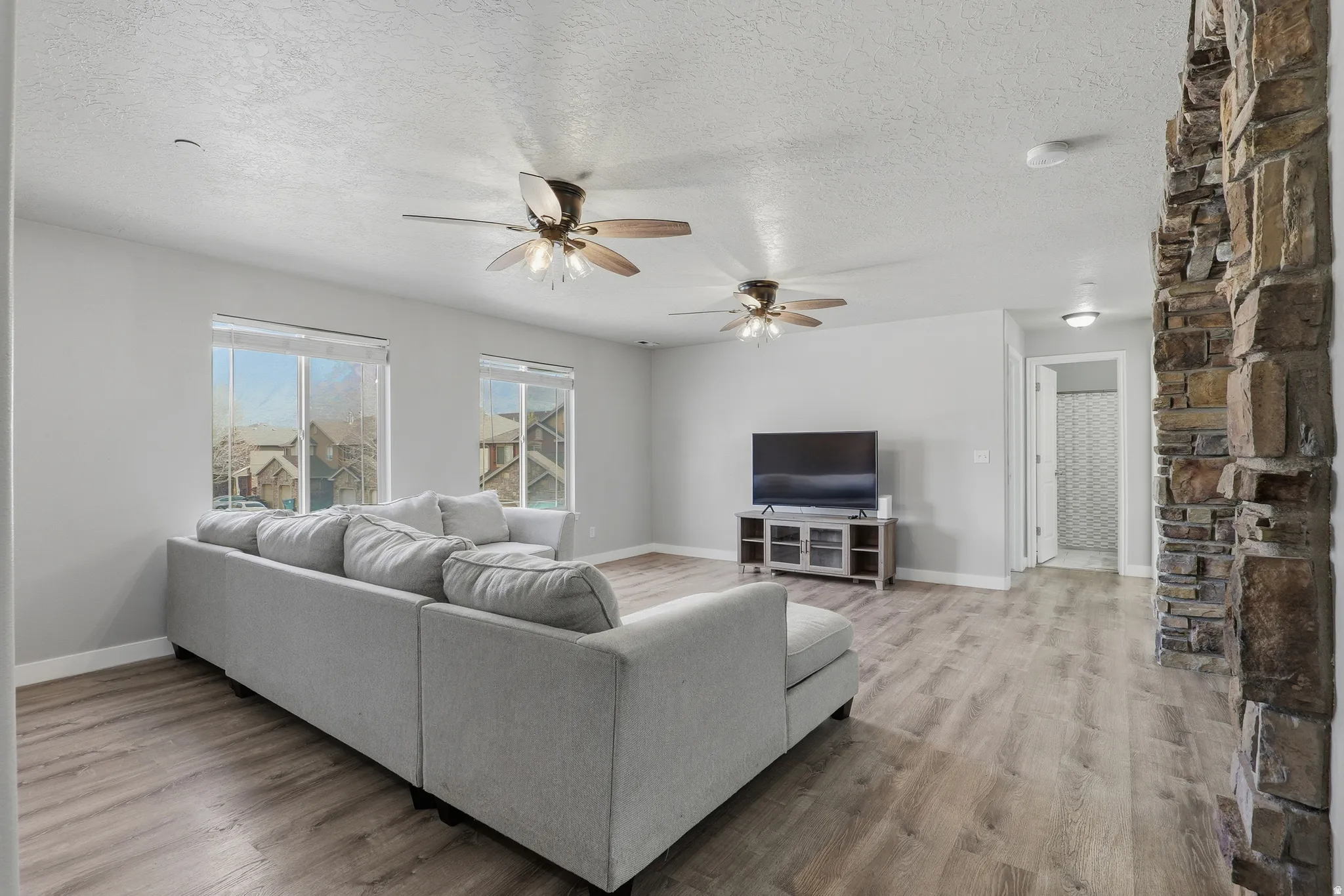 Spacious living area featuring wood-finish flooring, a stone-clad architectural element, multiple ceiling fans with integrated lighting, and large windows