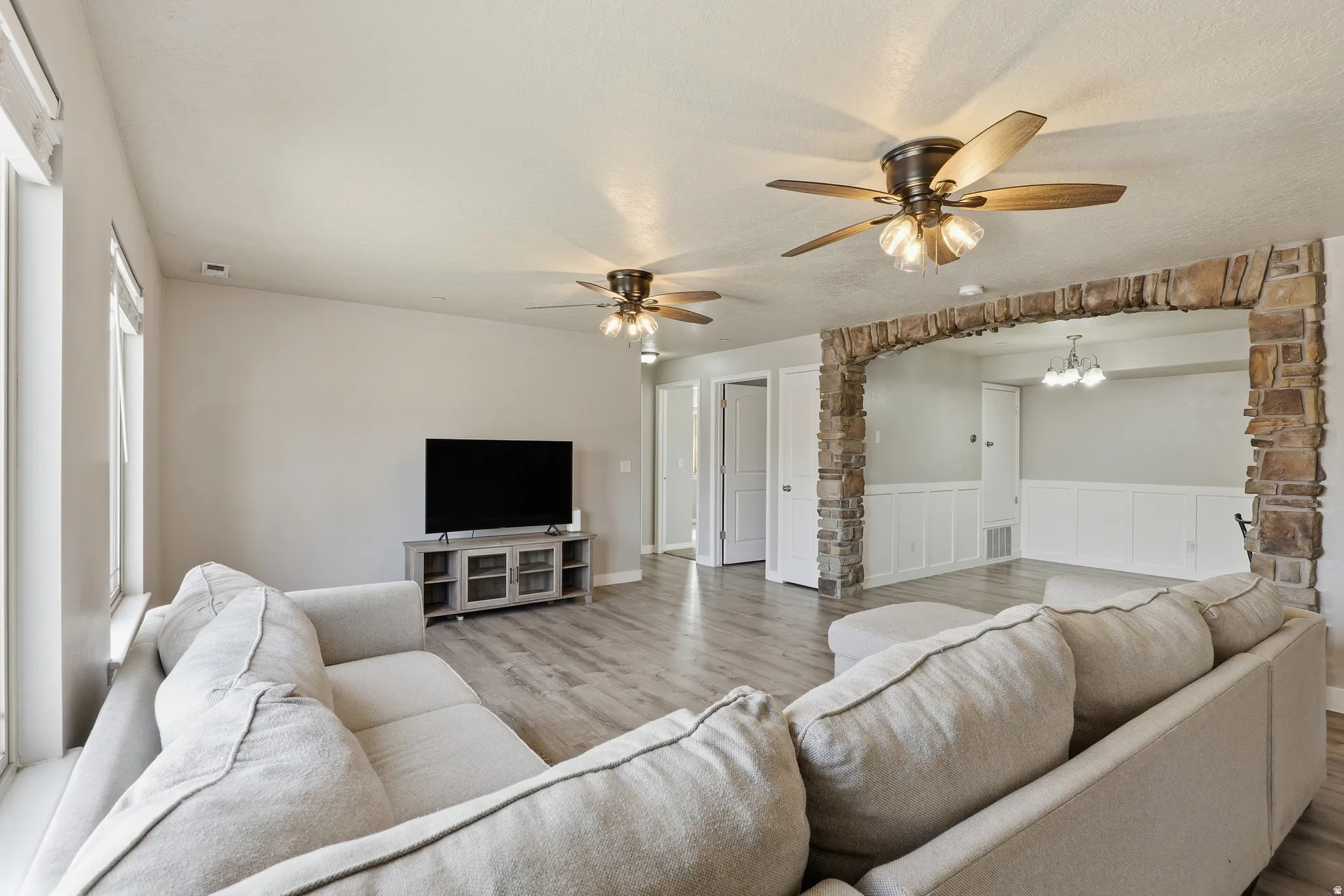Expansive living area featuring wood-finish flooring, a stone archway, multiple ceiling fans with integrated lighting, and transom windows