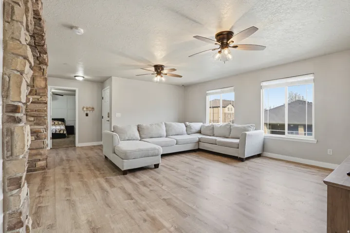 Spacious living area showcasing wood-finish flooring, a light gray upholstered sectional with chaise, dual ceiling fans with integrated lighting, large windows with city views, and a striking stacked-stone fireplace accent wall