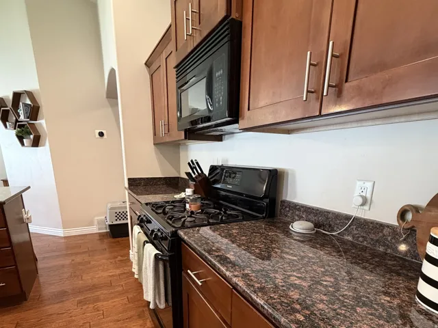 Wood-finish flooring, ample wood cabinetry, dark stone countertops, black gas range, and a black built-in microwave