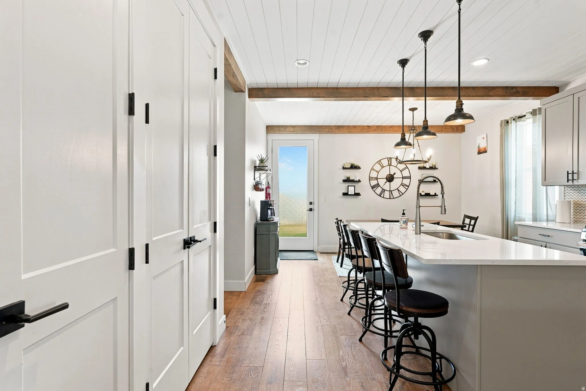 Wood-finish flooring, white panel doors with matte black hardware, exposed wood beams, and a kitchen island with pendant lighting