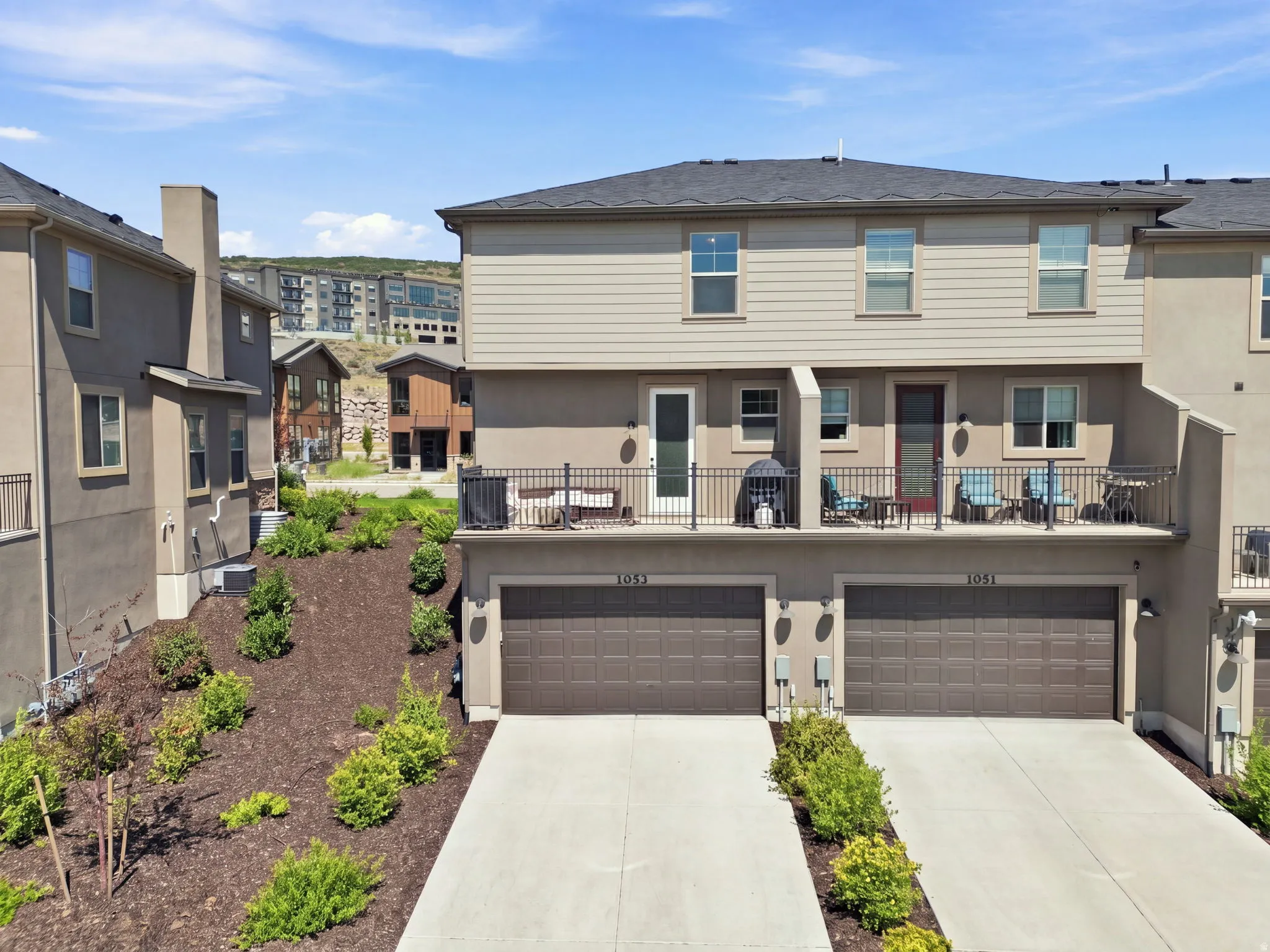 Two-car garage with individual doors, concrete driveways, upper-level balcony with metal railing, and a sloped, landscaped terrain
