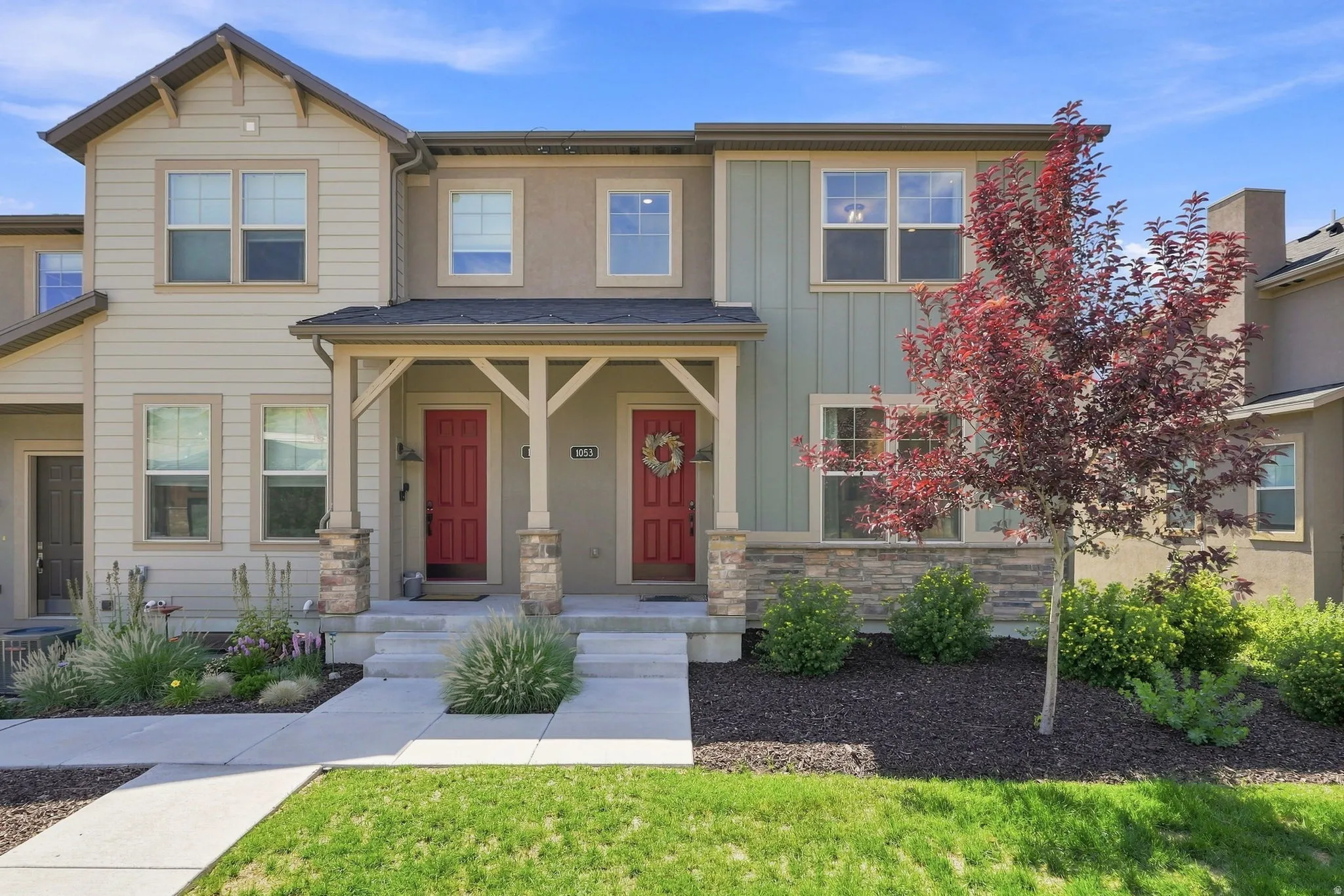 Multi-toned exterior with siding and stucco finishes, covered porch with stone-clad columns, red entry doors, and a landscaped front yard with a young tree