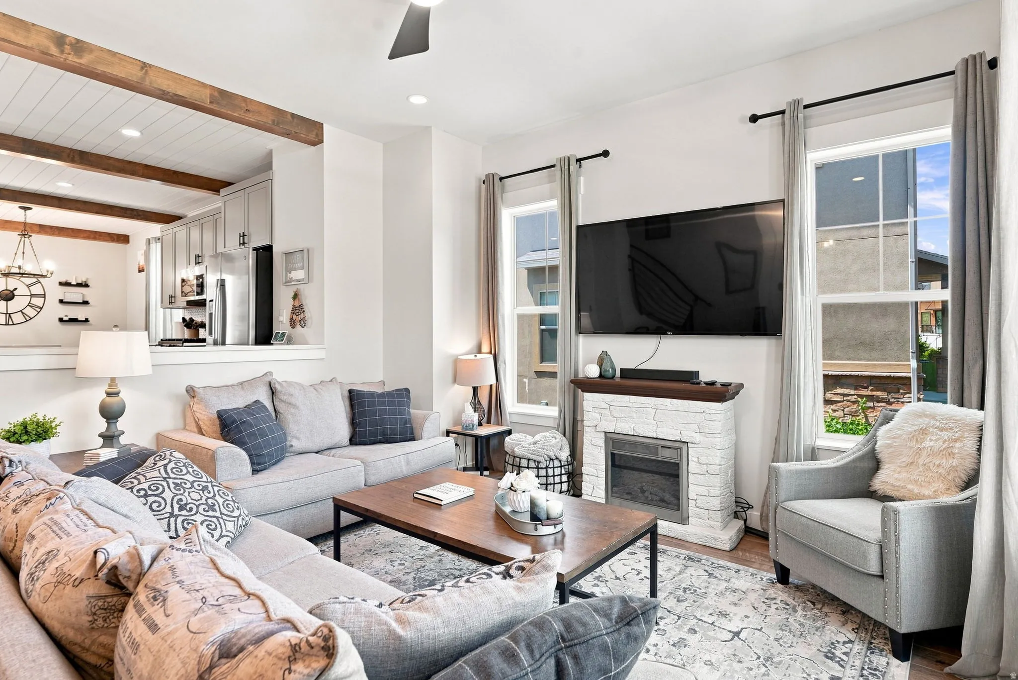 Built-in electric fireplace with stone-finish surround, recessed lighting, wood-finish ceiling beams, ceiling fan, and large windows